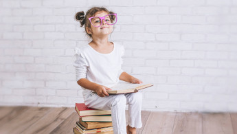 Ein Mädchen mit Brille sitzt auf einem Bücherstapel mit einem Buch in den Händen