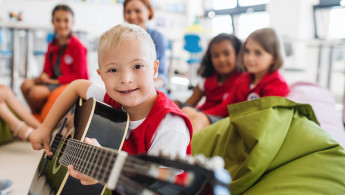 Fröhlicher Junge mit Downsyndrom spielt Gitarre in Mitten seiner Kita-Gruppe