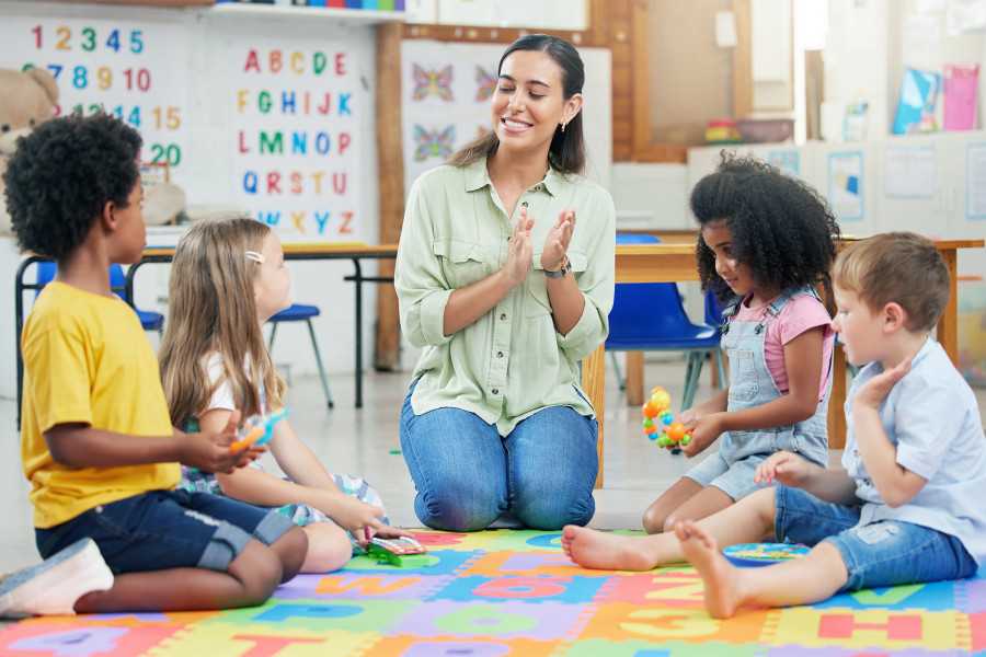 Erzieherin und Kinder sitzen mit Spielsachen in einem Kreis sie schauen sich gegenseitig freundlich an