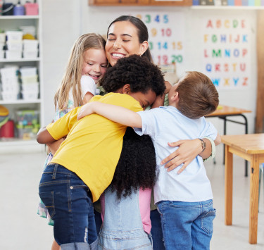 Erzieherin umarmt lachend mehrere Kinder in einer Kita-Gruppe