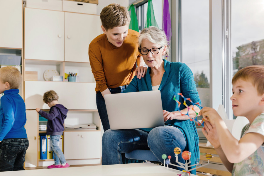 Zwei Erzieherinnen schauen – umgeben von Kindern – gemeinsam auf einen Laptop