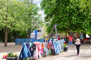 Jacken hängen auf dem Zaun der Kita Wirbelwind, dahinter spielen Kinder auf dem Spielplatz