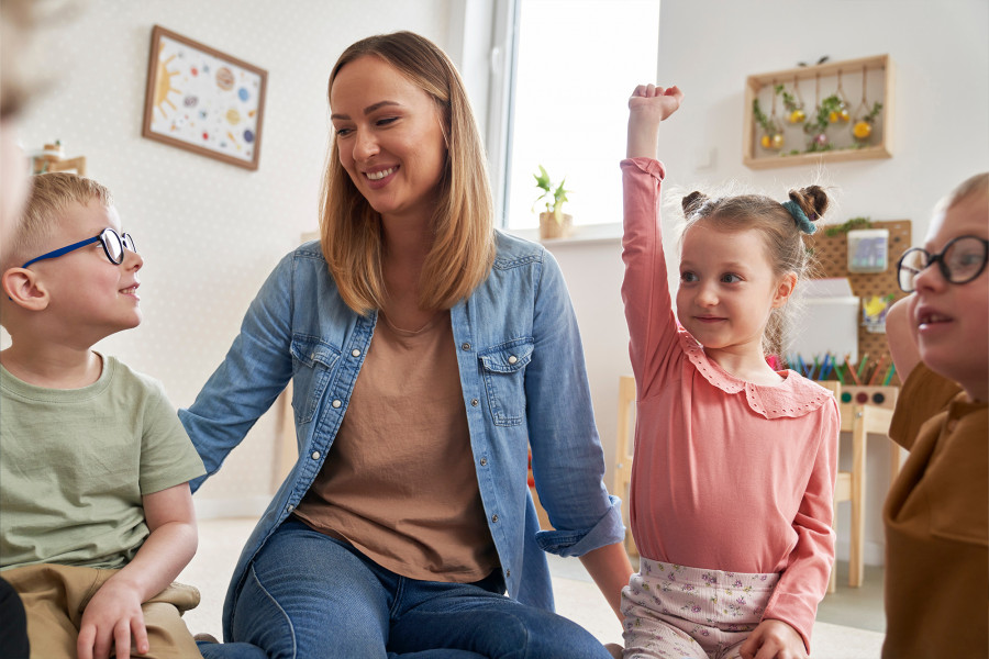 Erzieherin und Kinder sitzen lächelnd in einem Kreis – ein Kind meldet sich